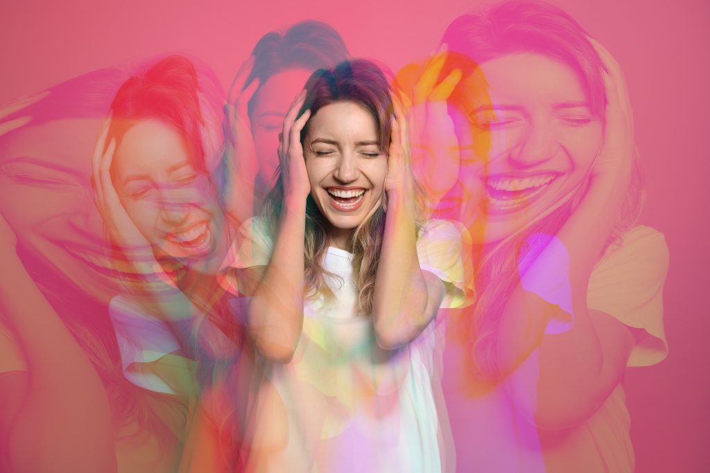 A joyful woman with long hair smiles broadly, holding her head, surrounded by colorful, blurred duplicates of herself.