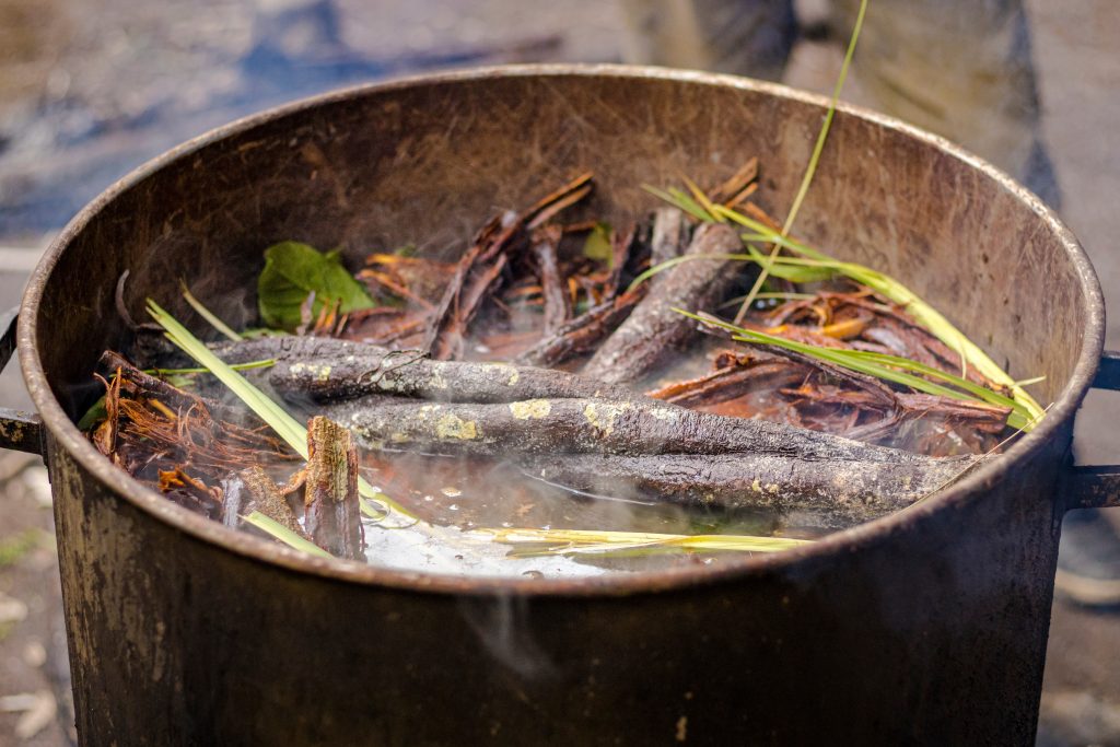 A large pot filled with boiling water, sticks, and leaves, emitting steam in an outdoor setting.