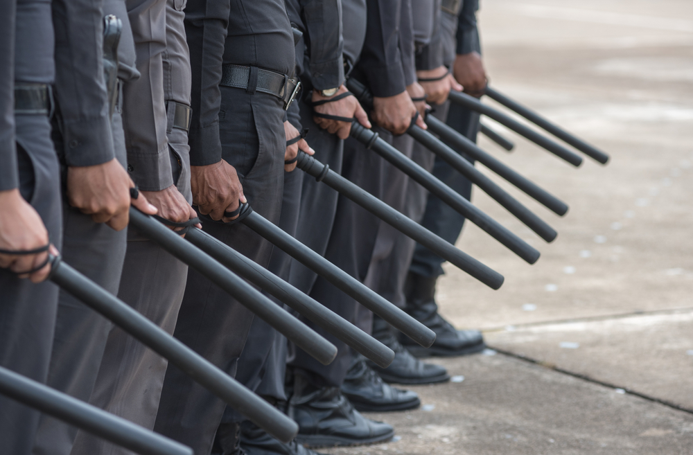 Line of uniformed officers holding batons, standing in formation on a concrete surface.