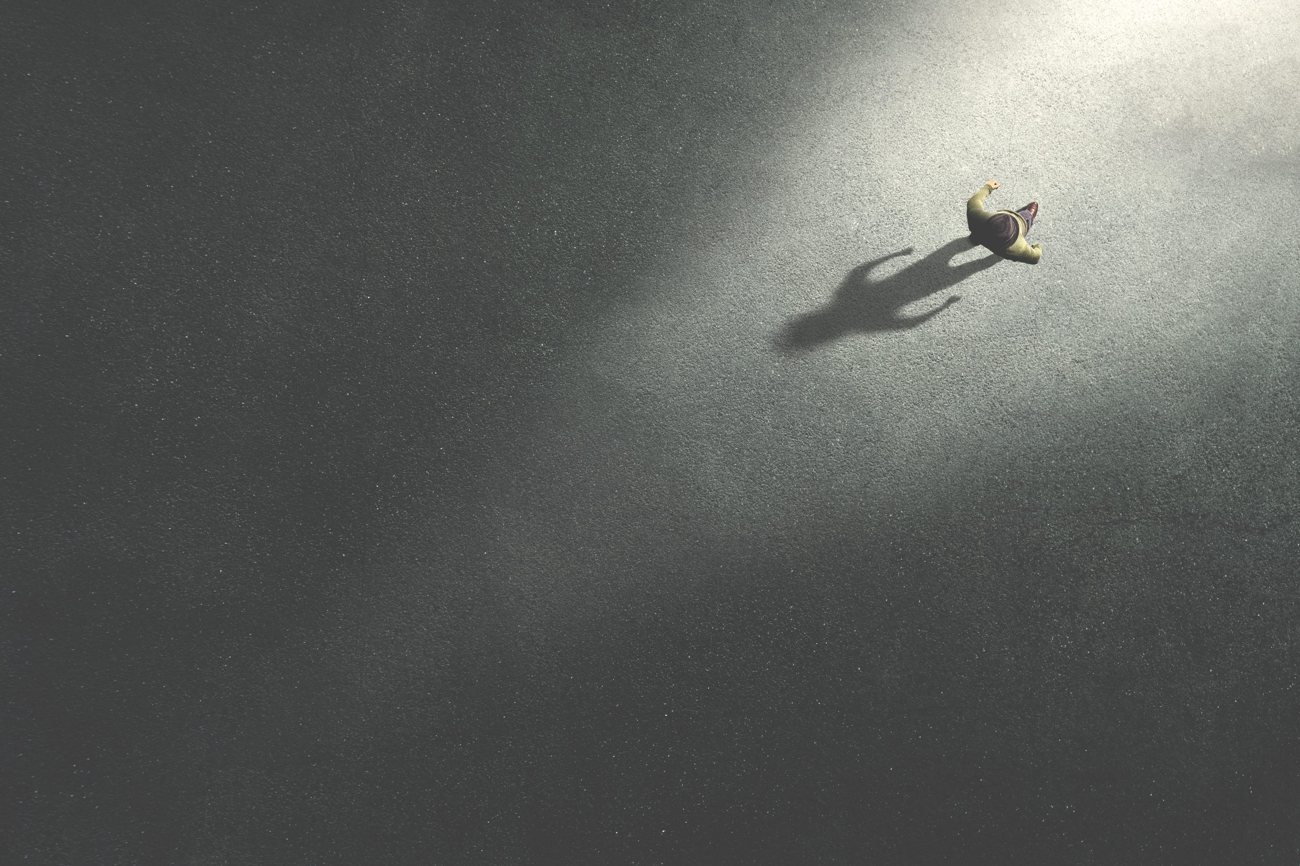 A lone figure stands on a dark surface, illuminated by a beam of light, casting a long shadow.