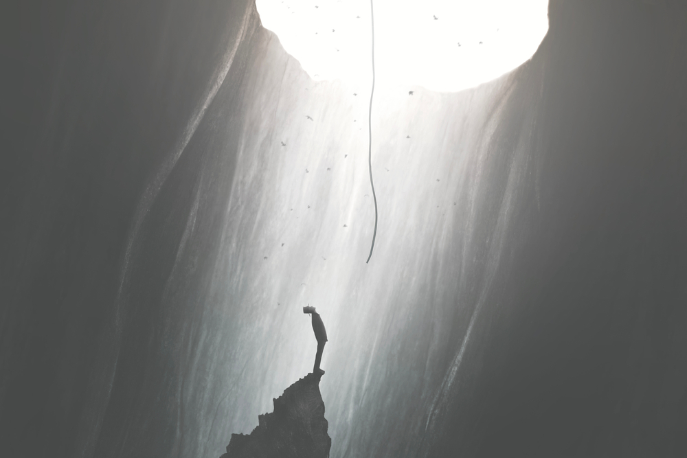 A lone figure stands on a rocky outcrop, gazing up at a bright light streaming down from above.