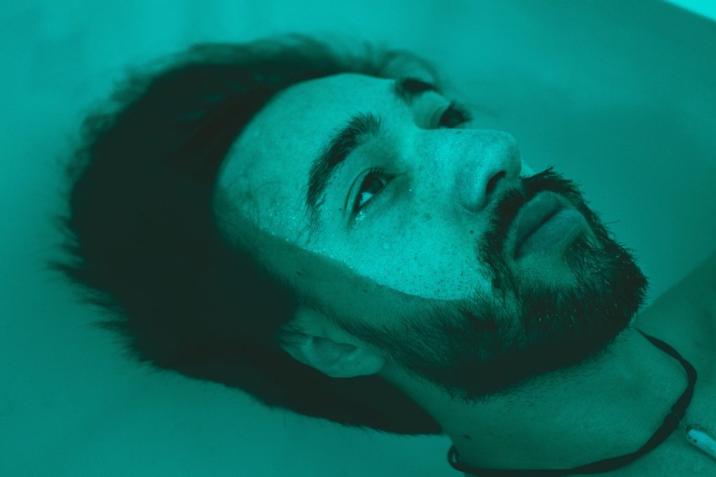 A man with a beard floats in water, his face illuminated in a teal hue, droplets glistening on his skin.