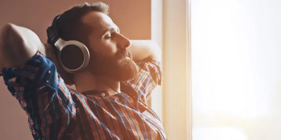 Man with a beard relaxing with headphones, eyes closed, enjoying music by a sunlit window.