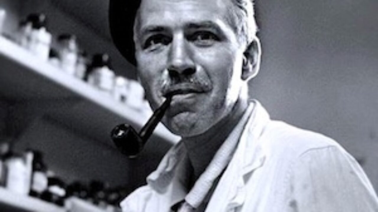 A man in a beret and lab coat smiles while holding a pipe, surrounded by shelves of bottles.