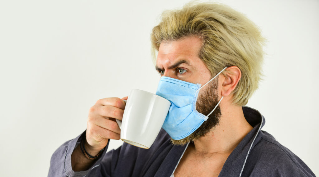 Man with blonde hair and beard wearing a blue mask, holding a white mug, looking thoughtfully to the side.