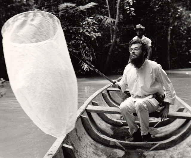 A man in a boat holds a fishing net, surrounded by lush jungle and another figure in the background.