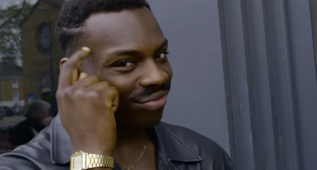 A man with a confident smile and slicked-back hair gestures to his head, suggesting cleverness or insight.