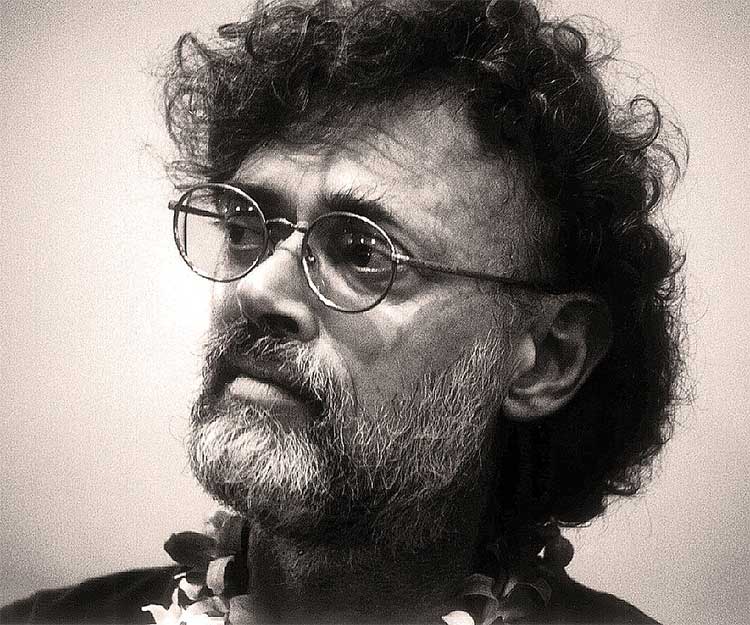 A man with curly hair and glasses gazes thoughtfully, wearing a floral necklace against a soft-focus background.