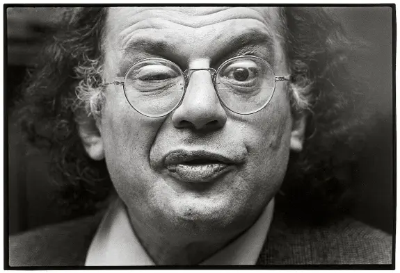 A man with curly hair and glasses makes a playful expression, winking and puckering his lips.