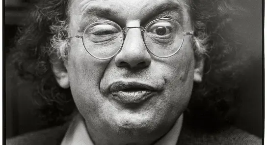 A man with curly hair and glasses makes a playful expression, winking and puckering his lips.