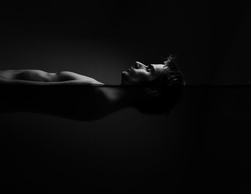 A man floats serenely in dark water, his profile reflected beneath the surface in a monochrome setting.
