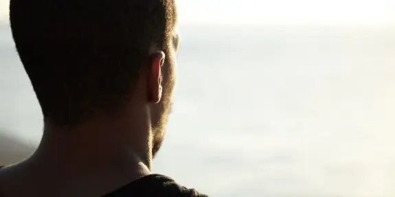 A man gazes thoughtfully at the ocean, sunlight reflecting on the water in the background.