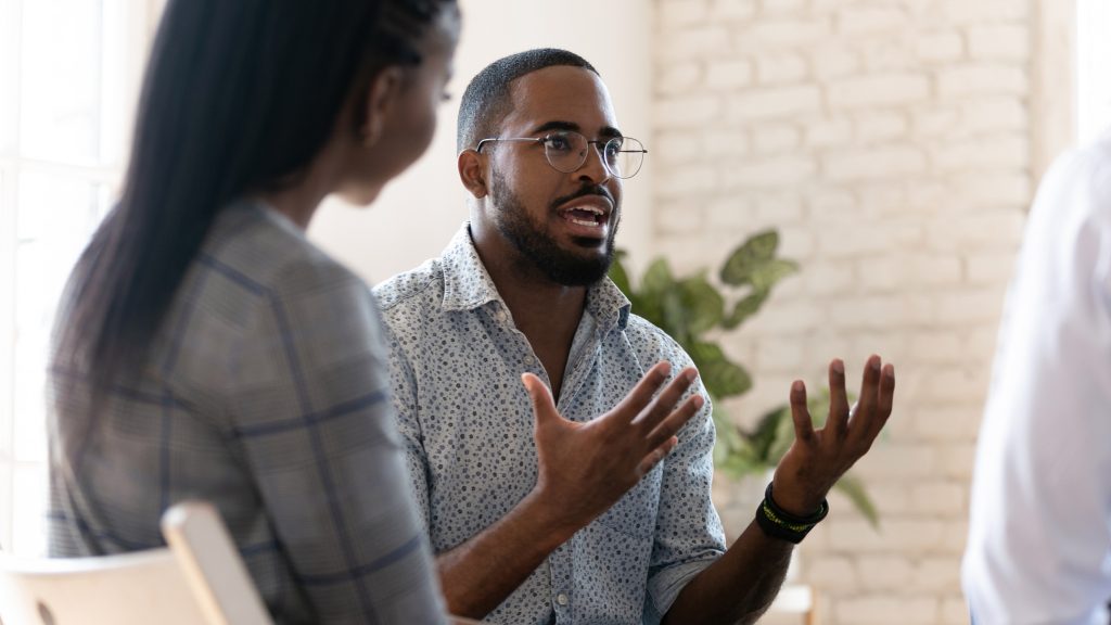 A man in glasses passionately discusses ideas with two people in a bright, modern room.