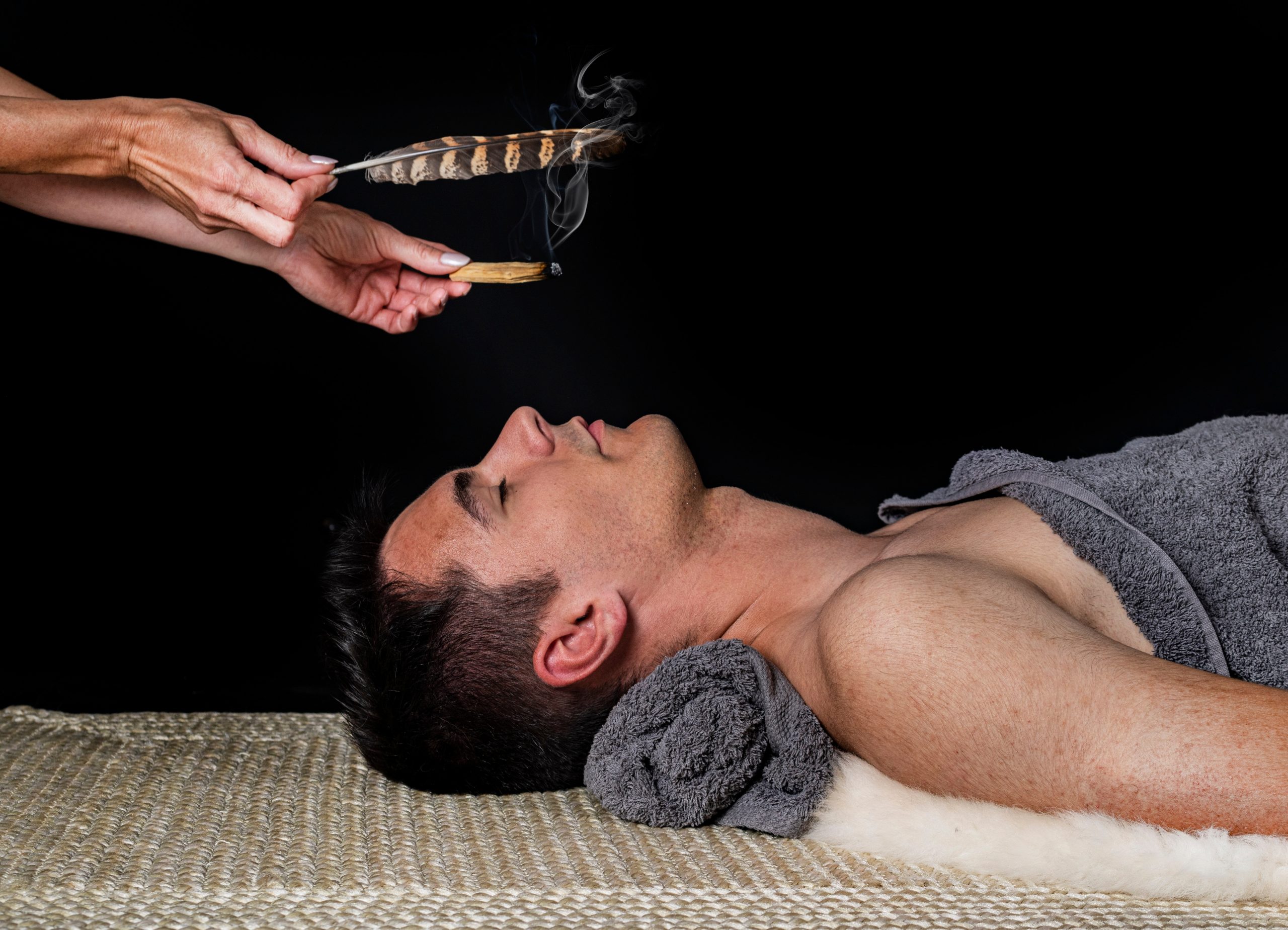 A man lies on a mat, eyes closed, as smoke from burning sage and feathers is waved above him.