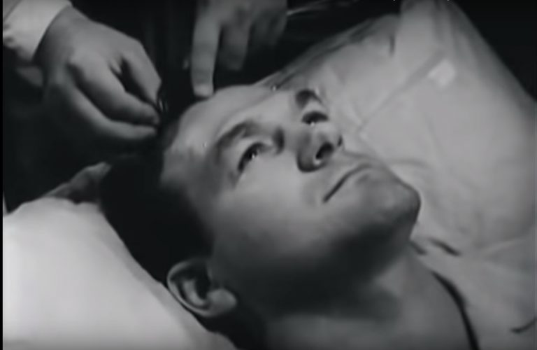 A man lies on a pillow, eyes closed, as hands adjust something in his hair.