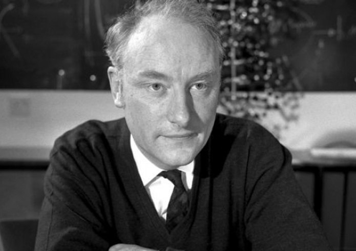 A man with light hair and a serious expression sits at a table, wearing a dark sweater and tie, against a blurred background.