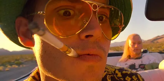 A man in oversized sunglasses and a green hat smokes a cigarette, with a woman sitting in the background.