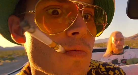 A man in oversized sunglasses and a green hat smokes a cigarette, with a woman sitting in the background.