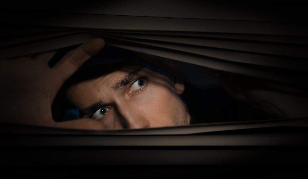 A man peeks through partially closed curtains, eyes wide with curiosity and apprehension in dim lighting.