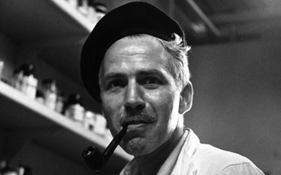 Man with a pipe and beret smiles, surrounded by shelves of jars and bottles in a workshop setting.
