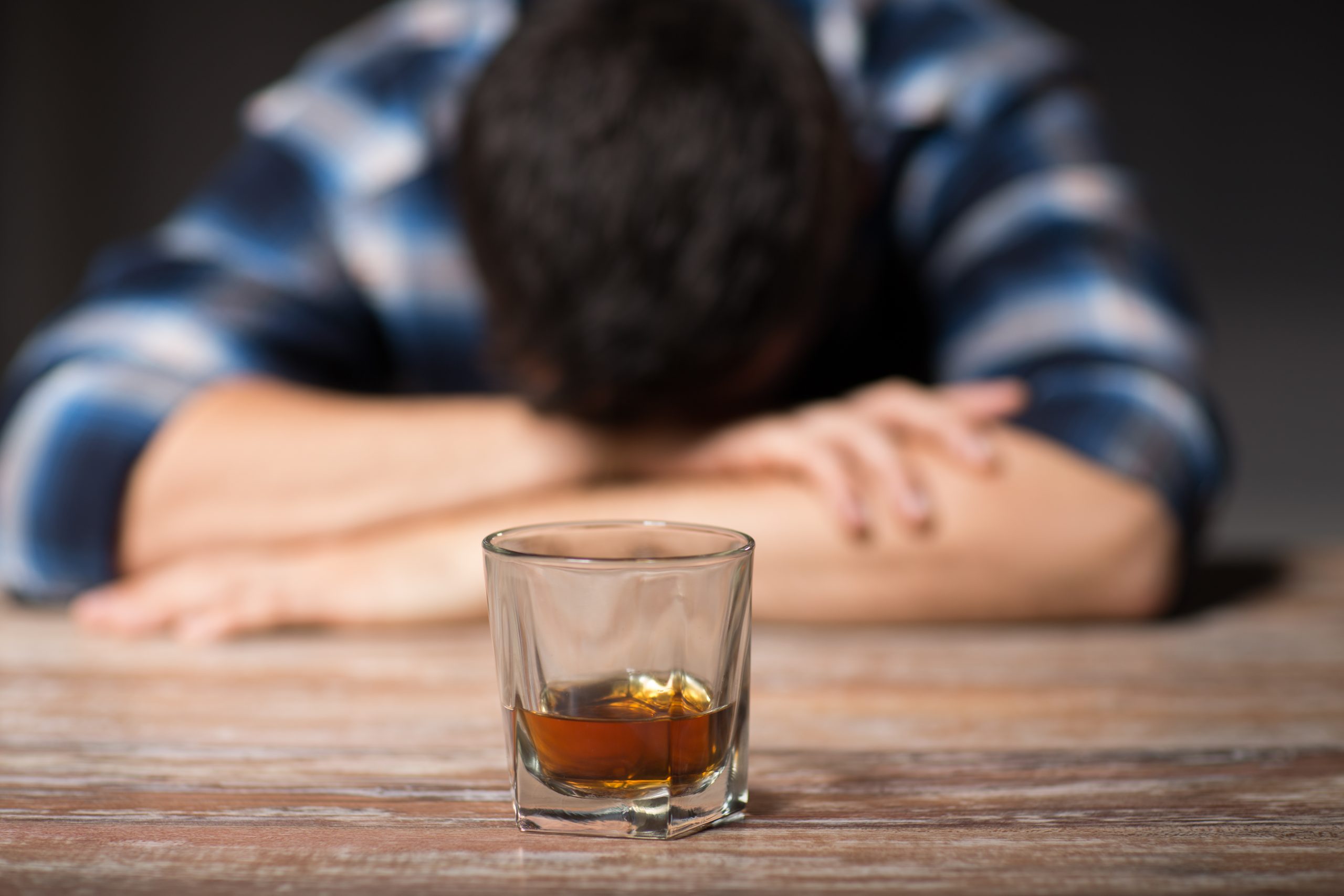 A man in a plaid shirt rests his head on crossed arms, with a glass of whiskey on a wooden table in front of him.