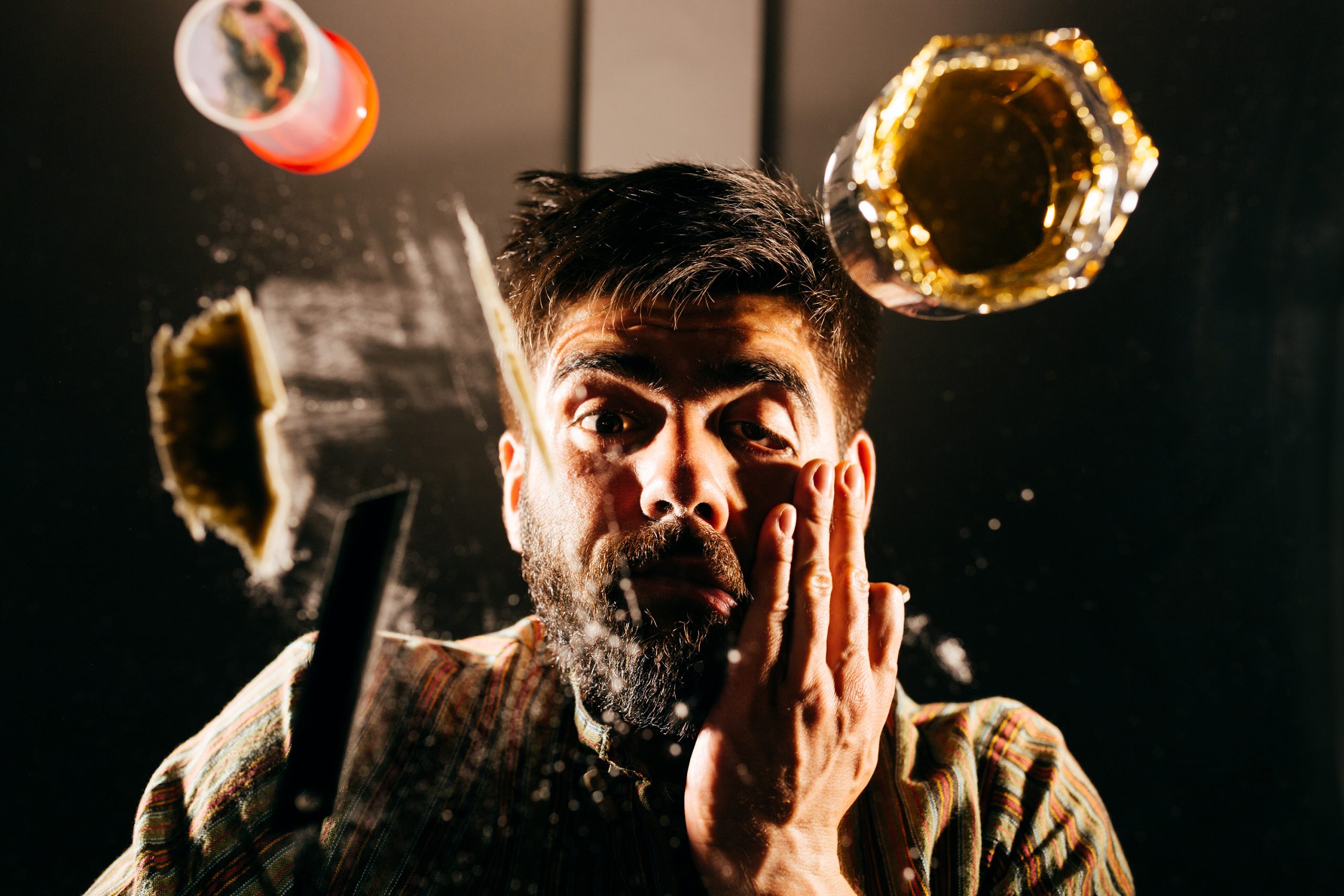 A man with a shocked expression, surrounded by flying objects, including a glass and a plate, against a dark background.