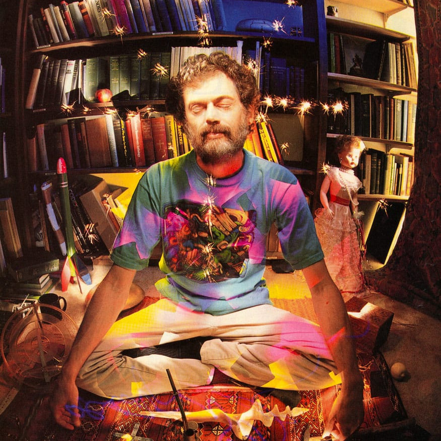 A man sits cross-legged in a colorful room filled with books, surrounded by swirling lights and a doll in the background.