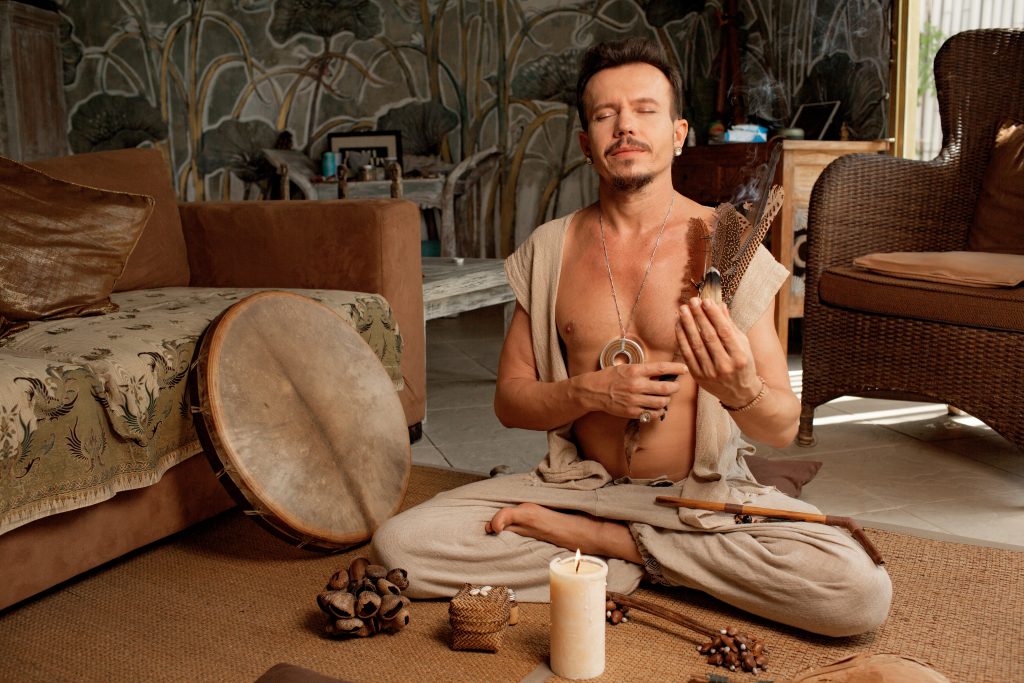 A man sits cross-legged in a serene room, holding a feathered instrument, surrounded by natural decor and soft lighting.