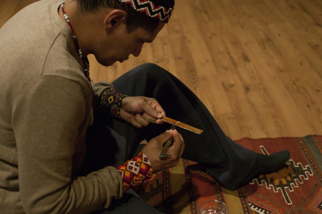A man sits on a patterned rug, carefully crafting a wooden object, wearing traditional attire with colorful embroidery.