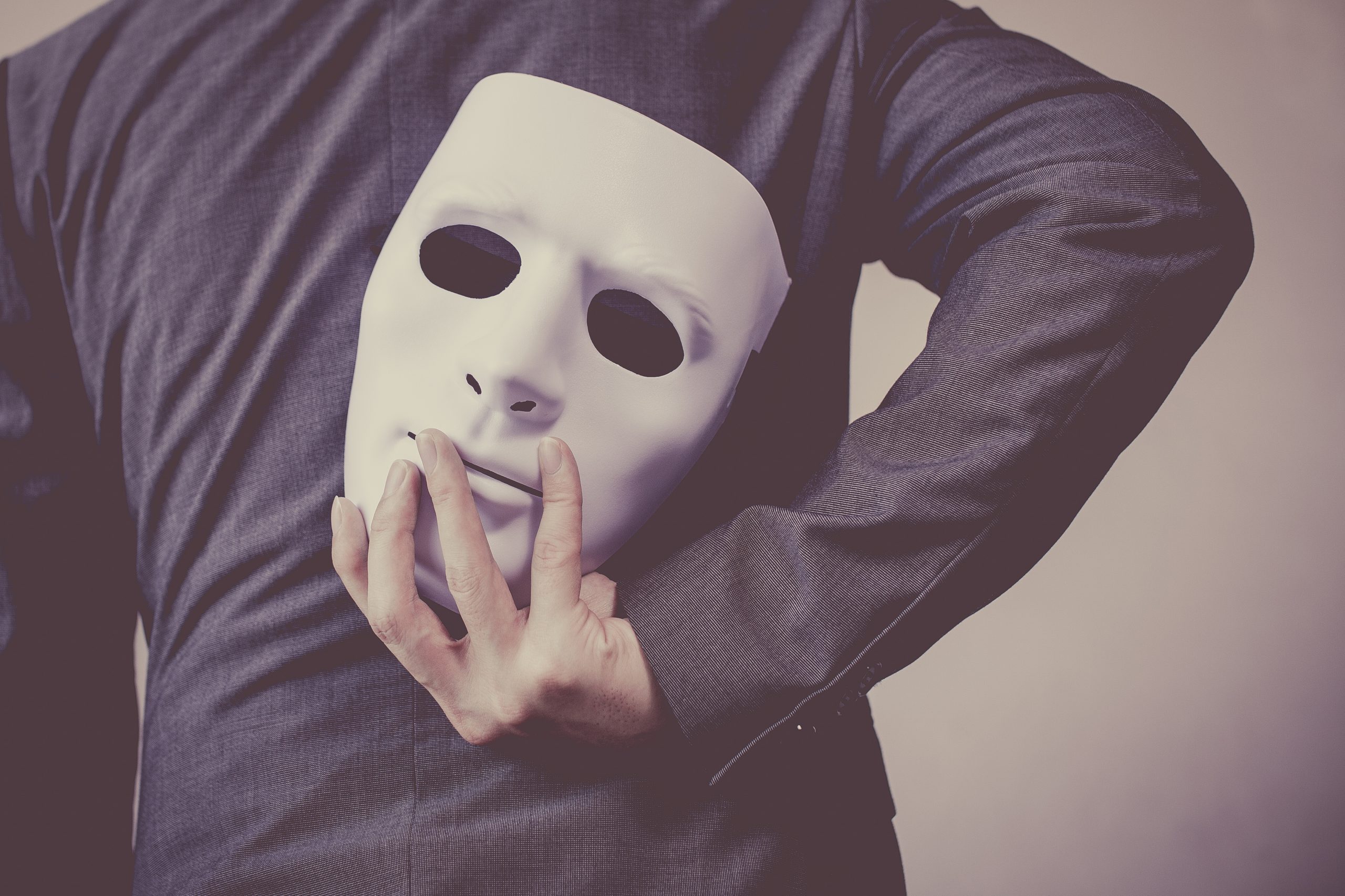 A man in a suit holds a white mask behind his back, suggesting deception or hidden identity.
