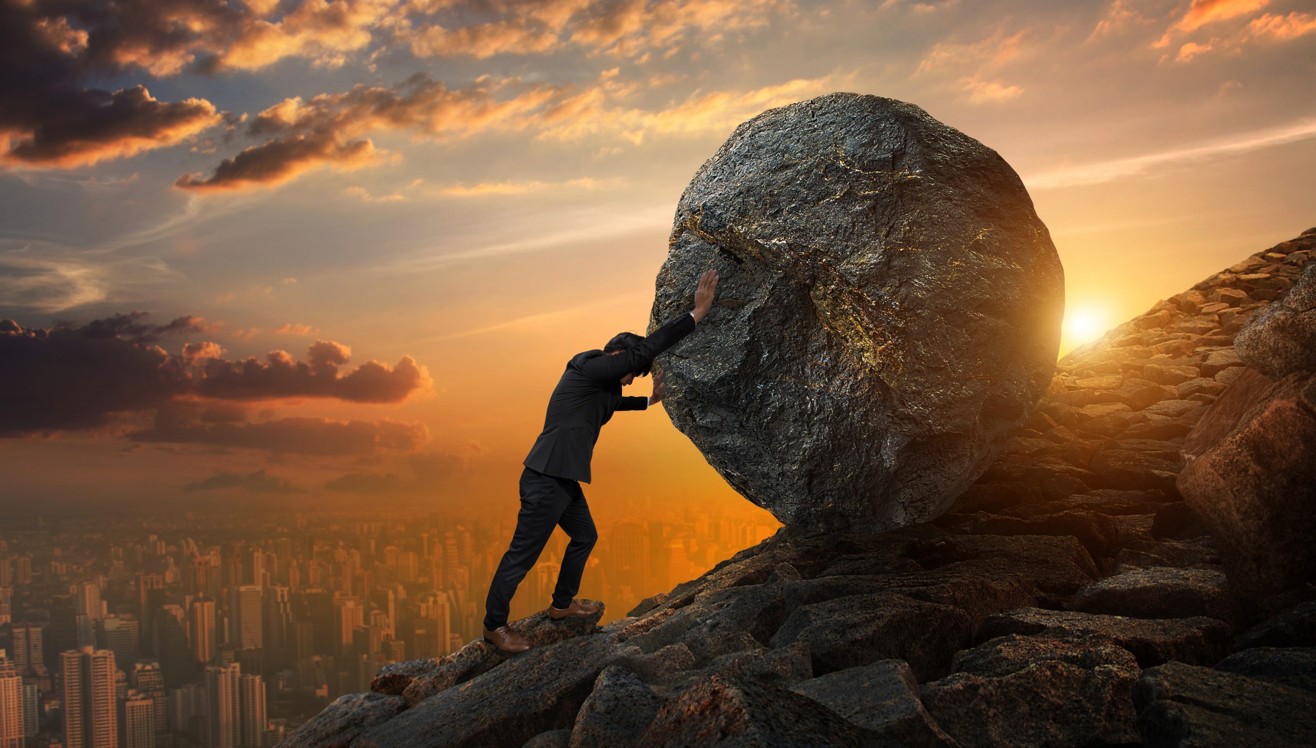 A man in a suit struggles to push a large boulder uphill against a sunset backdrop over a city skyline.