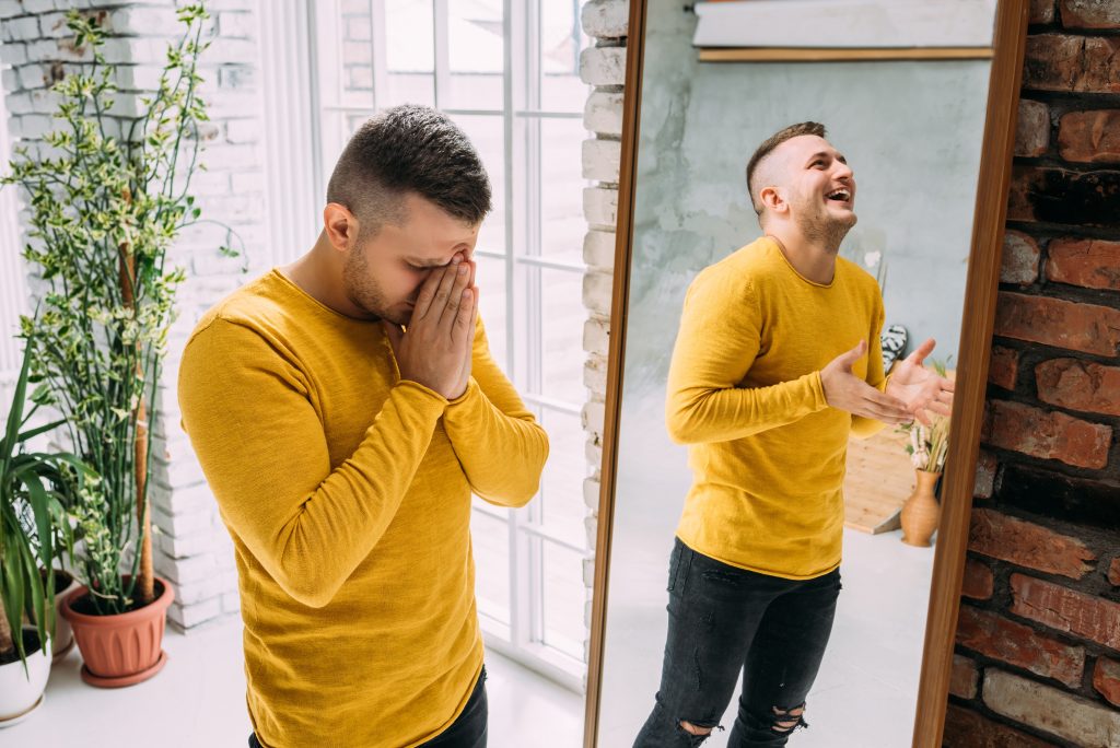 Man in a yellow sweater stands by a mirror, one hand on his face, while laughing joyfully. Indoor plants in the background.