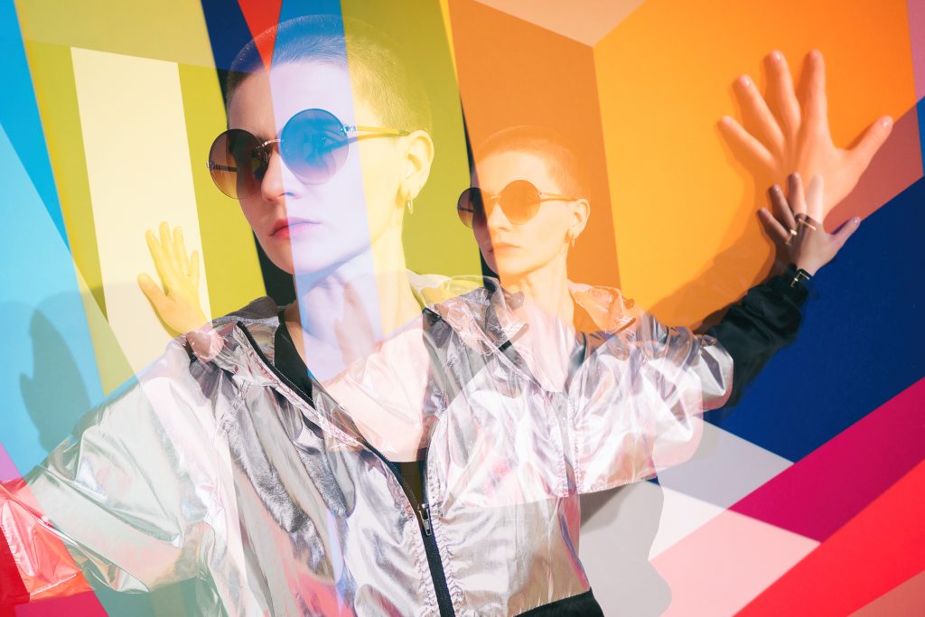 Model in oversized sunglasses poses against a vibrant, multicolored geometric background, creating a striking visual effect.