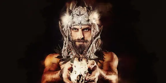 A muscular man with long hair and a horned headdress holds a skull, surrounded by dark, dramatic lighting.