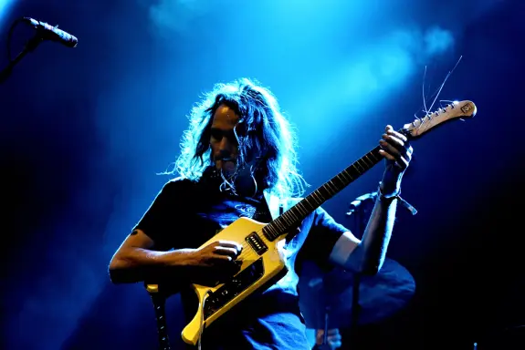 Musician with long hair plays a yellow guitar under dramatic blue lighting on stage.