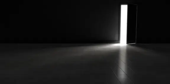 A partially open door emits bright light into a dark, empty room, casting a long shadow on the tiled floor.