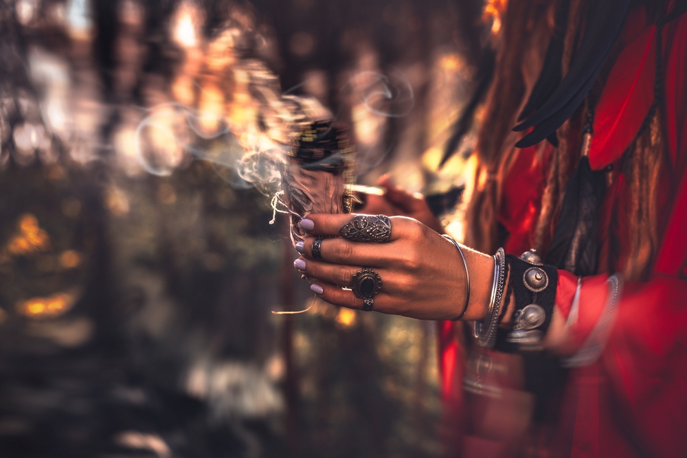 A person with adorned hands holds a bundle of herbs, surrounded by a blurred forest background and wisps of smoke.