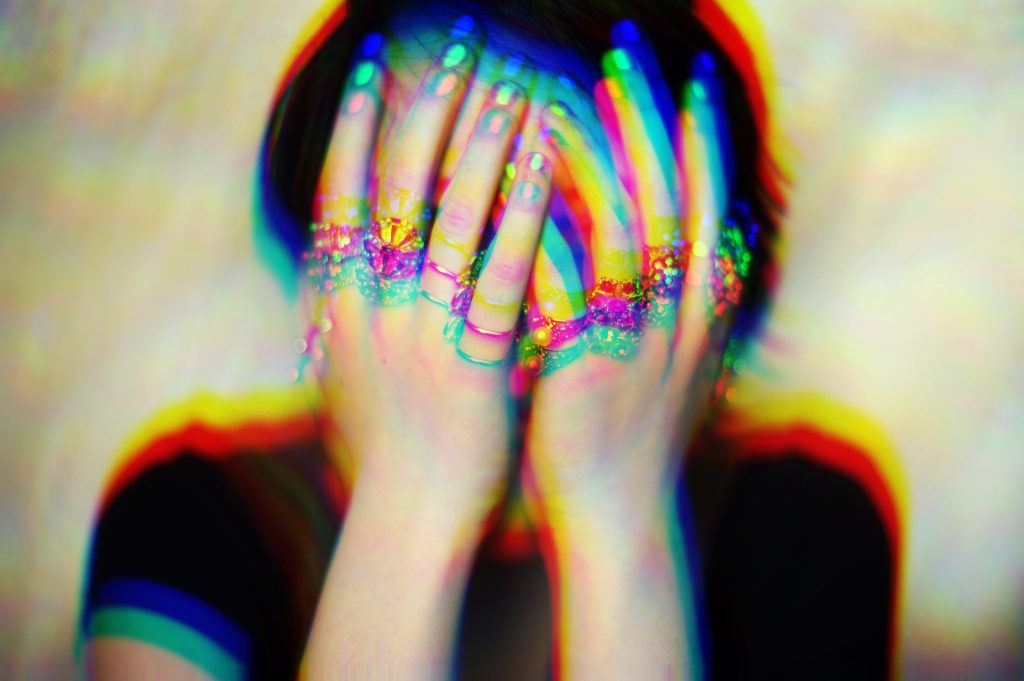 A person with colorful, distorted lighting covers their face, adorned with multiple rings on their fingers.