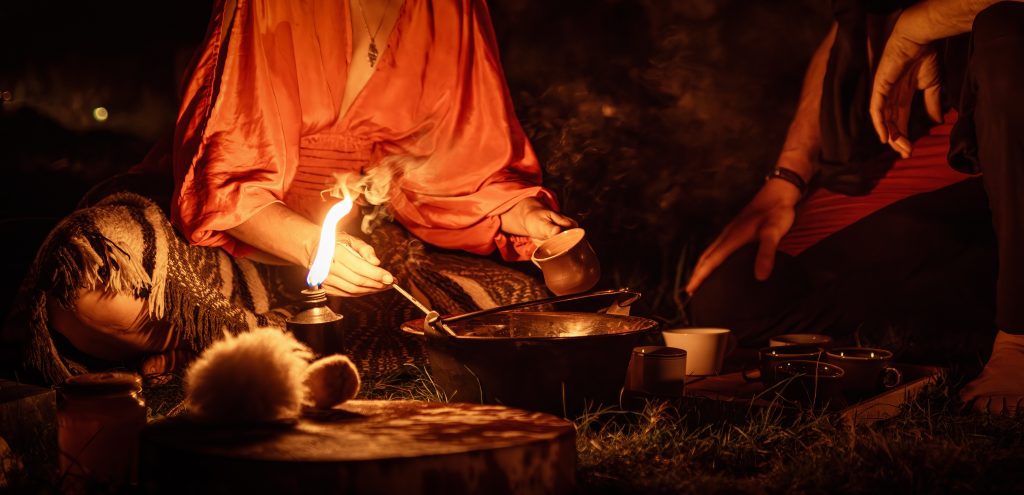 A person in a flowing red robe pours from a pot, surrounded by flickering candlelight and smoke in a dark, outdoor setting.