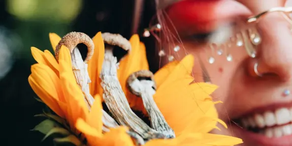 A person with glasses smiles while holding a sunflower adorned with mushrooms.