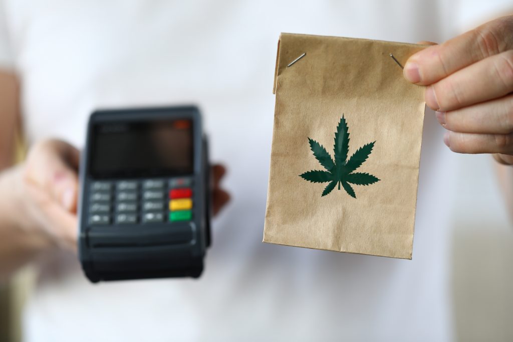 A person holds a brown paper bag with a cannabis leaf design while displaying a payment terminal.