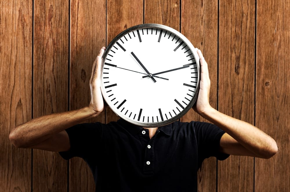 A person holds a large clock in front of their face against a wooden wall, emphasizing the passage of time.