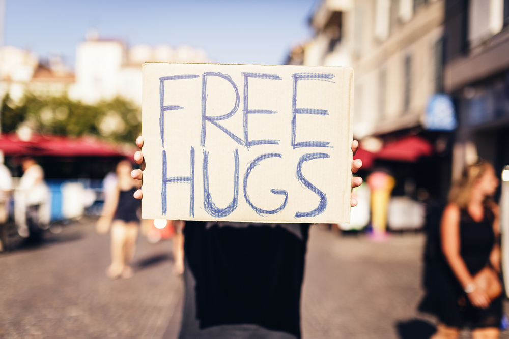 A person holds a large sign reading "FREE HUGS" in a bustling outdoor setting.