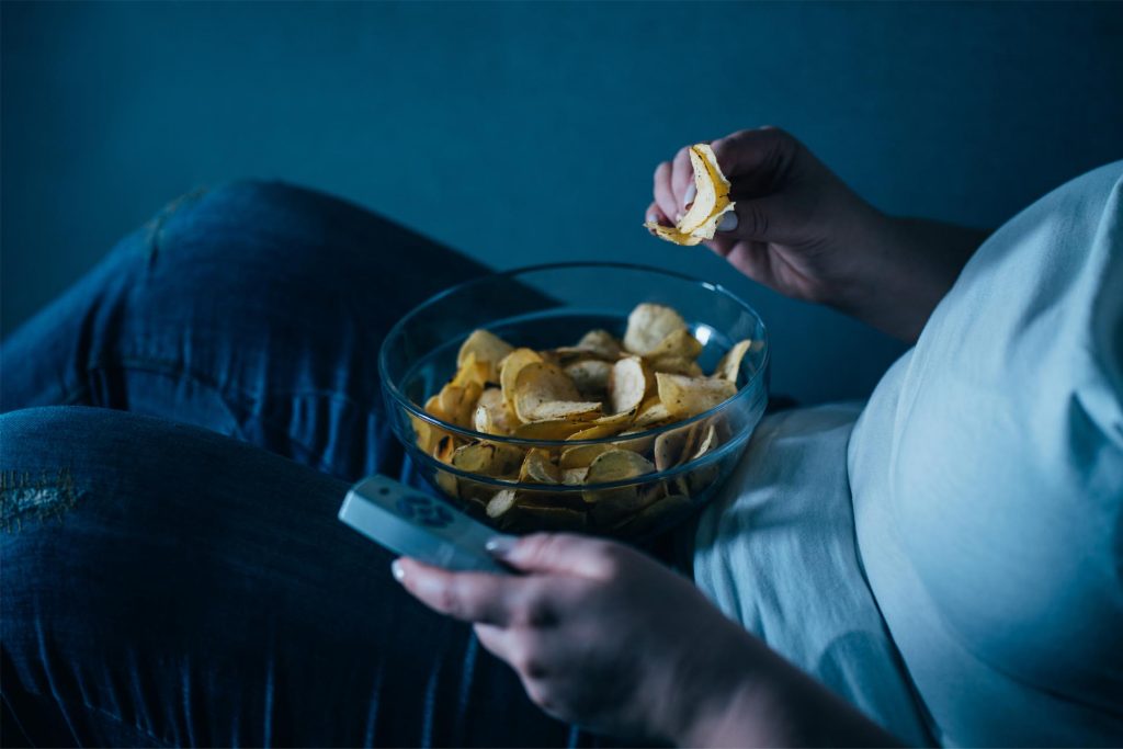 A person holds a potato chip above a bowl while sitting with a remote control in hand, relaxed on a couch.
