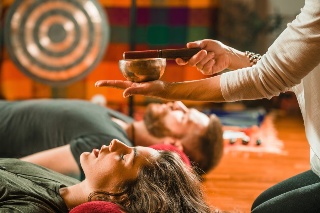 A person holds a singing bowl above a woman's face while a man lies relaxed in the background, surrounded by colorful text...