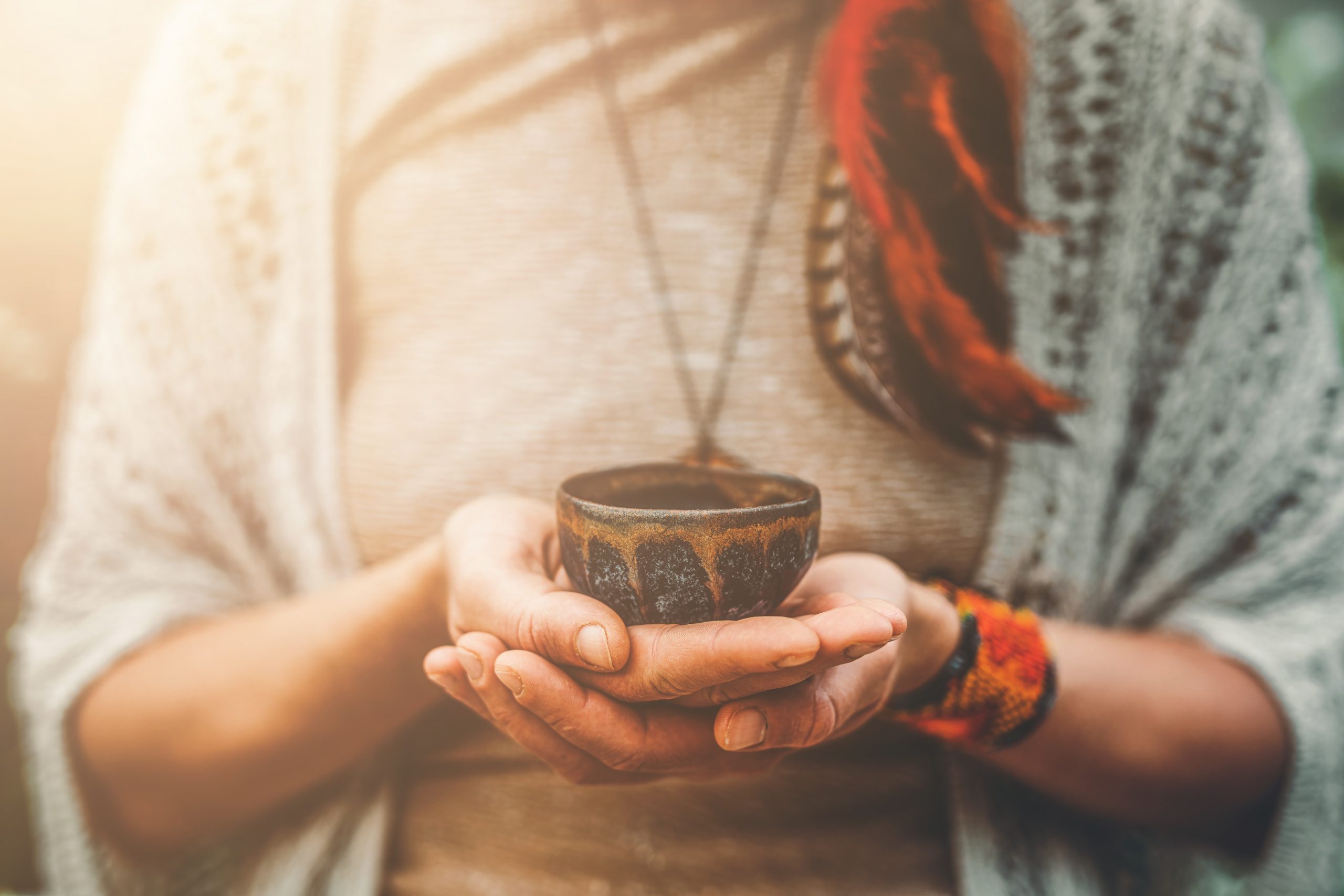 A person holds a small, ornate bowl in their hands, wearing a cozy sweater and adorned with feathers and colorful bracelets.