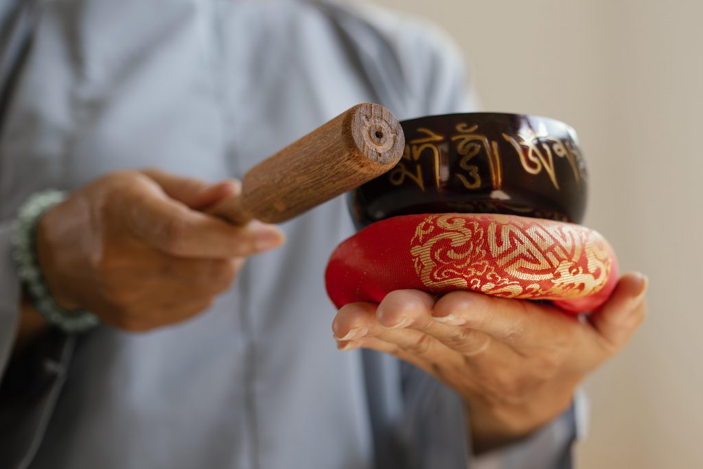 A person holds two singing bowls, one black with inscriptions and one red with intricate patterns, alongside a wooden mallet.