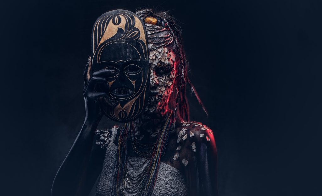A person with intricate body paint and a decorative mask, surrounded by dark tones, exuding a mystical aura.