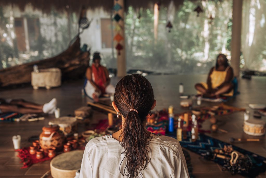 A person with long hair in a ponytail sits in a circle with others, surrounded by colorful objects and natural decor.