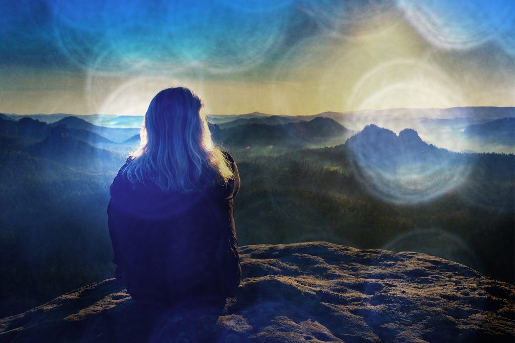 A person with long hair sits on a rock, gazing at a misty mountain landscape under a colorful sky.